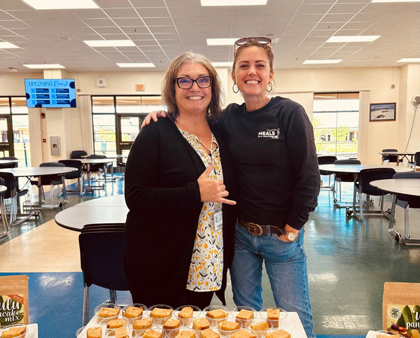 Two women posing together in a school cafeteria with food items on tables.